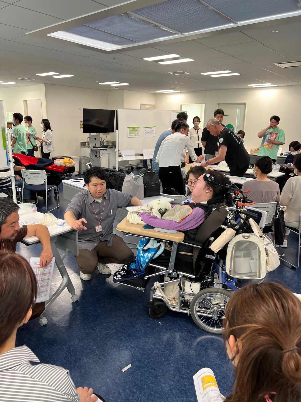 車いすの参加者を含む人たちが同じ輪になり、器材を前に話し合うセミナーの場面